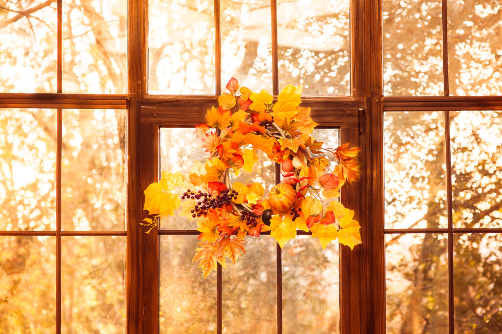 Autumn wreath entwined with leaves, garlic, berries, pumpkins, mushroom, hanging on the wooden window during sunset.