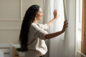 Perfect Morning Concept. Profile side view of smiling young woman opening window curtains at home to let the sunshine in, enjoying start of the new day, standing in modern bedroom alone