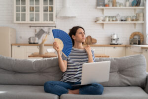 Tired millennial woman suffering from heatstroke flat without air-conditioner, waving blue fan, sitting on couch at home, working on laptop computer. Overheating, high temperature, hot summer weather.