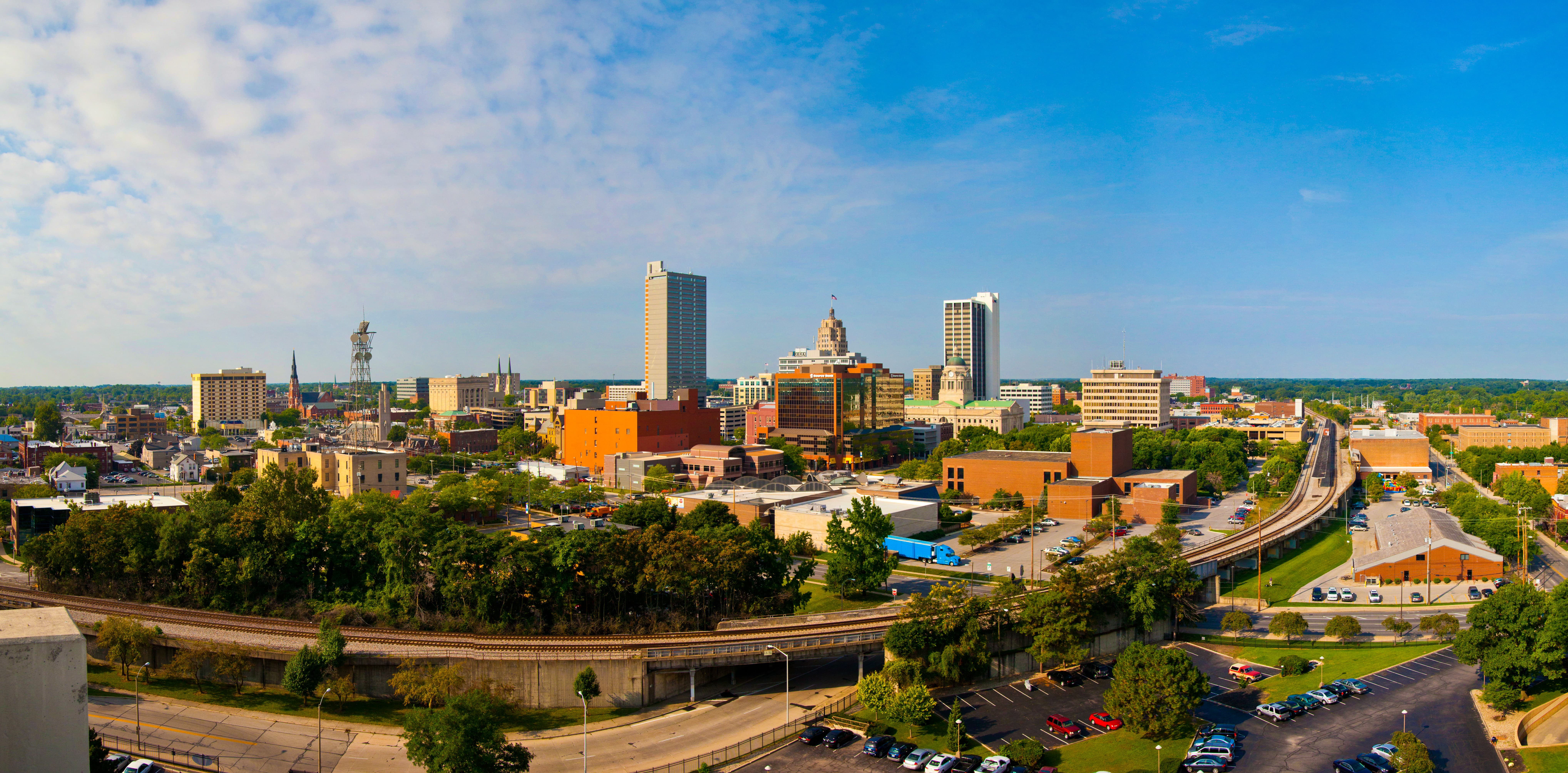 A picturesque city skyline with various buildings is nestled behind a lush green park with winding roads, under a dramatic sunset sky