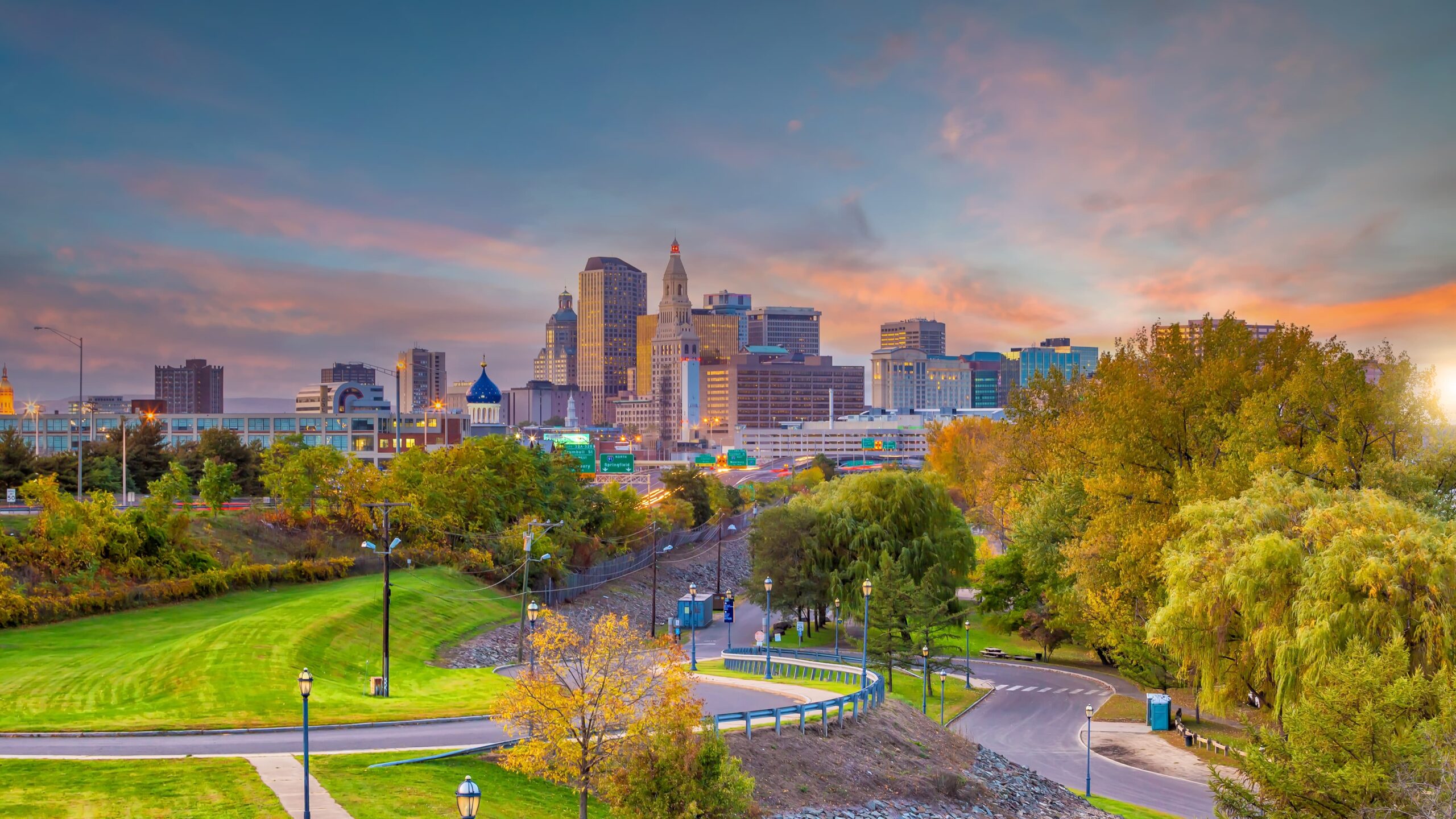 AdobeStock_415342552 An aerial view captures a diverse city landscape with tall buildings, suburban areas, a river, and green spaces under a cloudy sky
